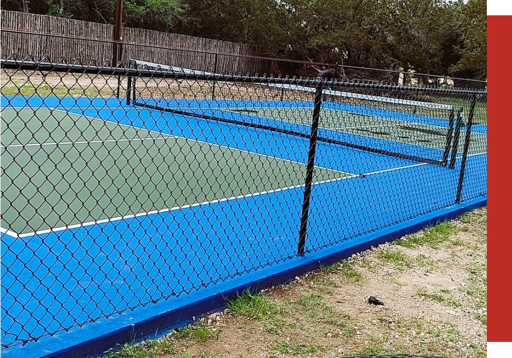 A fenced outdoor tennis court with blue and green playing surfaces, surrounded by grass and trees.