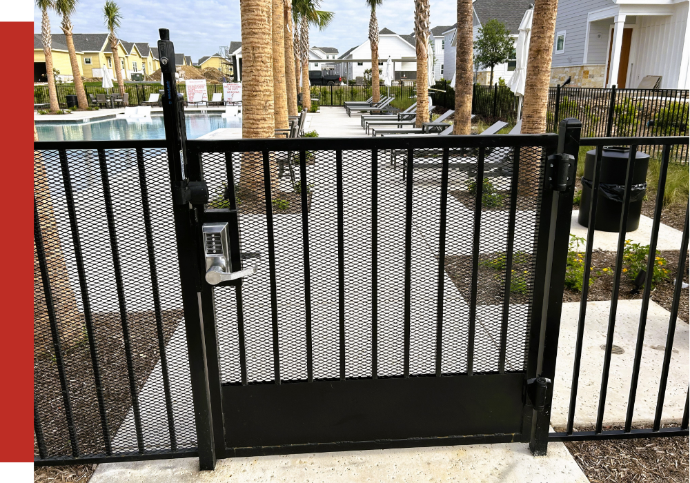A black metal gate with a keypad lock secures entry to a community outdoor pool area with lounge chairs, palm trees, and surrounding houses.