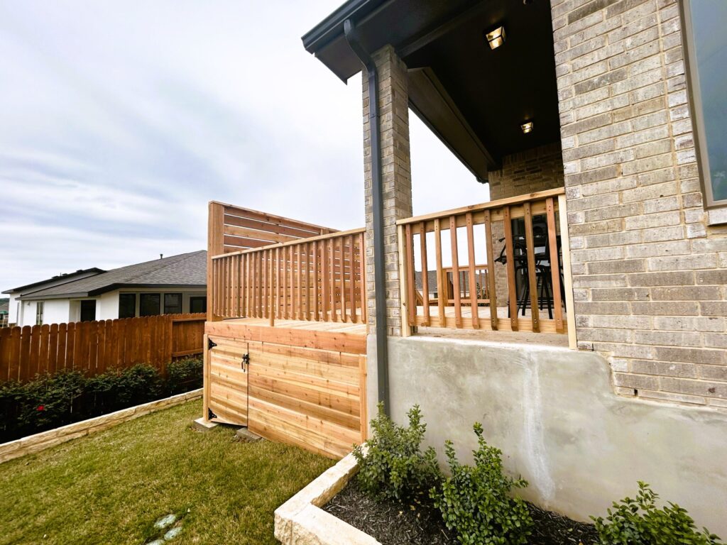 A wooden deck with railing and privacy screen is attached to a brick house, overlooking a lawn and garden bed with shrubs.