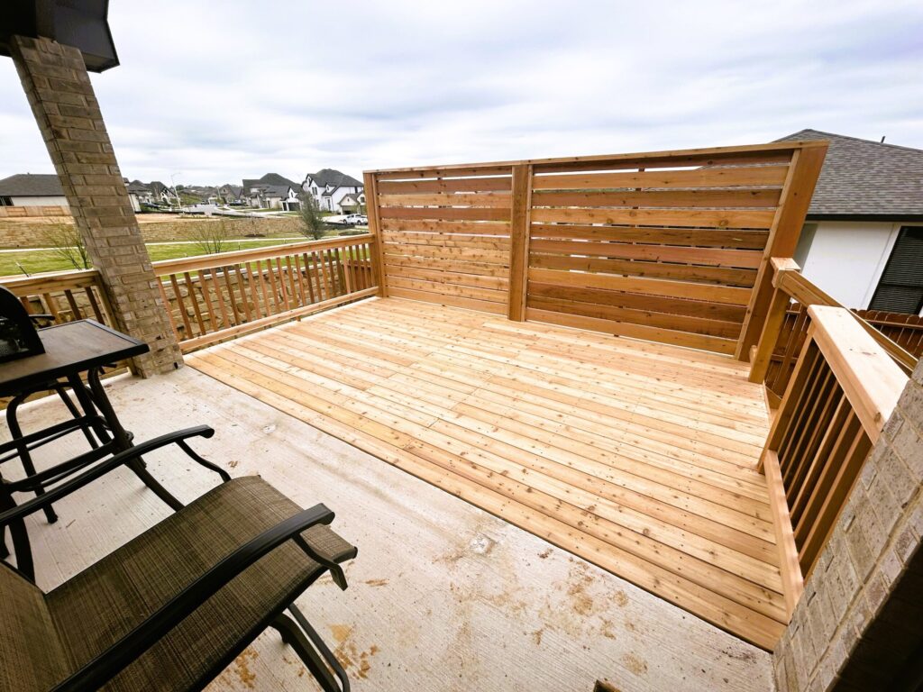 A wooden deck with a privacy fence and railing, attached to a house. Two patio chairs are placed on the concrete floor next to the deck. Suburban houses are visible in the background.