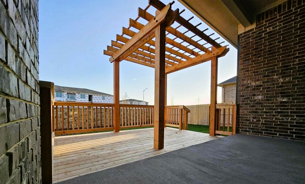 A wooden deck with a pergola, surrounded by a fence and brick walls, overlooking a suburban neighborhood under a clear sky.
