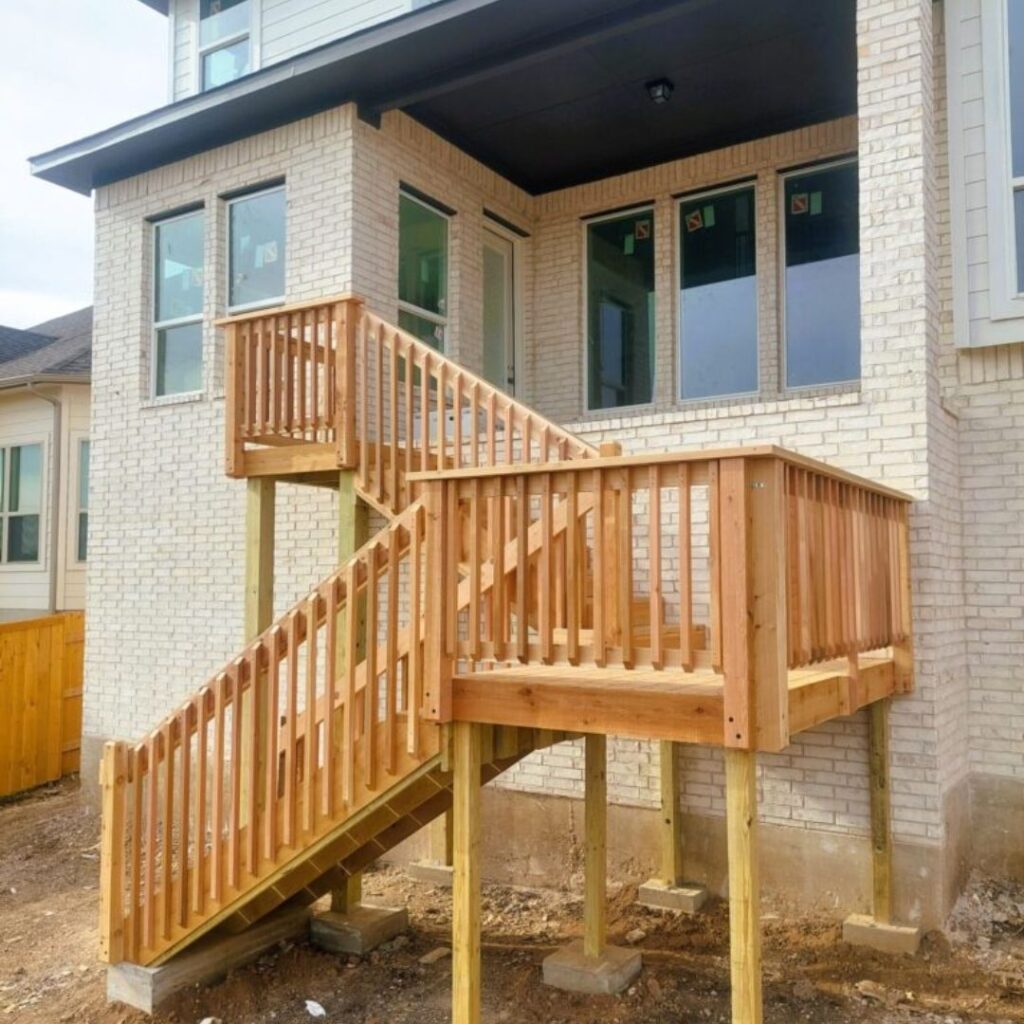 A newly built wooden deck with stairs and railings is attached to the back of a light brick house, elevated above bare soil.