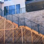 %Imagetitle% | Discount Fence Usa Outdoor wooden staircase with black metal railings and lattice paneling, attached to a stone wall of a modern building with large windows.