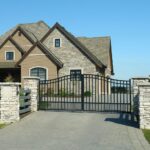 A modern house with stone and wood exterior is seen behind a closed black metal gate and stone pillars, with a paved driveway in front.