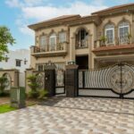 Two-story beige house with decorative railings, large windows, a tiled driveway, and ornate black and gold gates, surrounded by a well-kept lawn and trees.