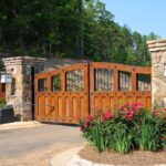 A wooden driveway gate with decorative ironwork is partially open, surrounded by stone pillars, flowering plants, and trees in the background.