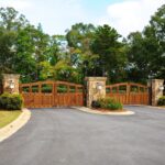 A large wooden driveway gate with stone pillars stands closed at the entrance to a property, surrounded by trees and greenery.