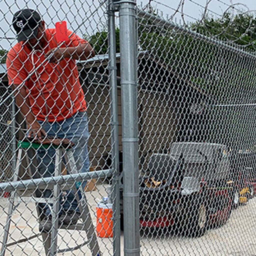 A man in a red shirt stands on a ladder to work on a chain-link fence topped with barbed wire; lawnmowers are visible behind the fence.