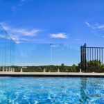 %Imagetitle% | Discount Fence Usa A swimming pool with a clear glass safety fence and metal railing overlooks a green landscape under a bright blue sky.