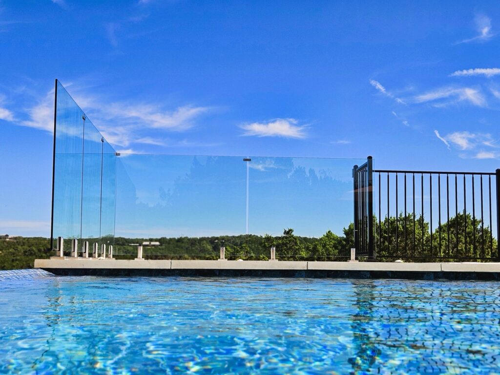 A swimming pool with a clear glass safety fence and metal railing overlooks a green landscape under a bright blue sky.
