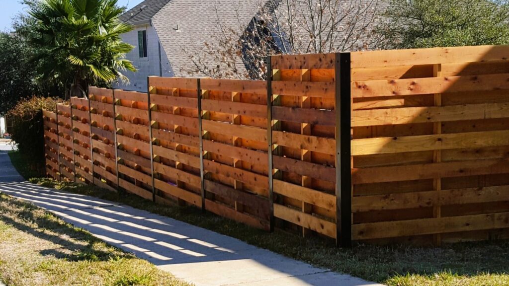 A wooden privacy fence with horizontal planks runs alongside a sidewalk, with grass and trees visible in the background.