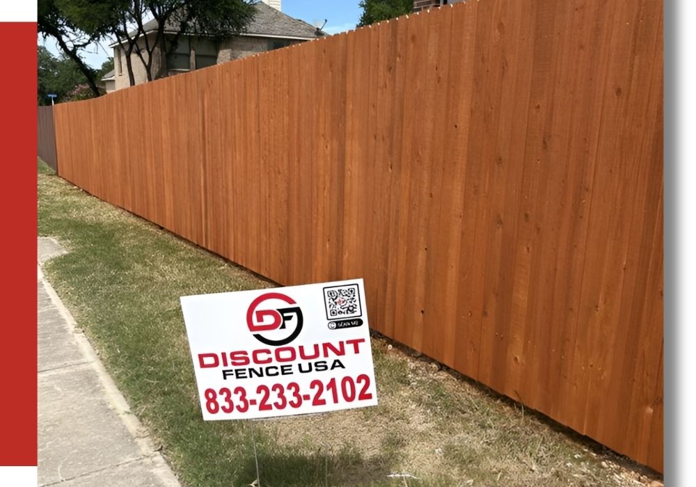 A tall wooden fence runs along a sidewalk with a “Discount Fence USA” sign and phone number posted on the grass in front.