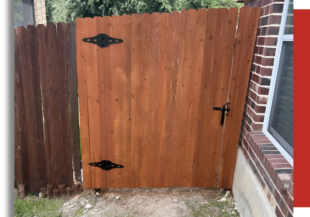 A wooden gate with black metal hinges and latch is attached between a brick house and a wooden fence.