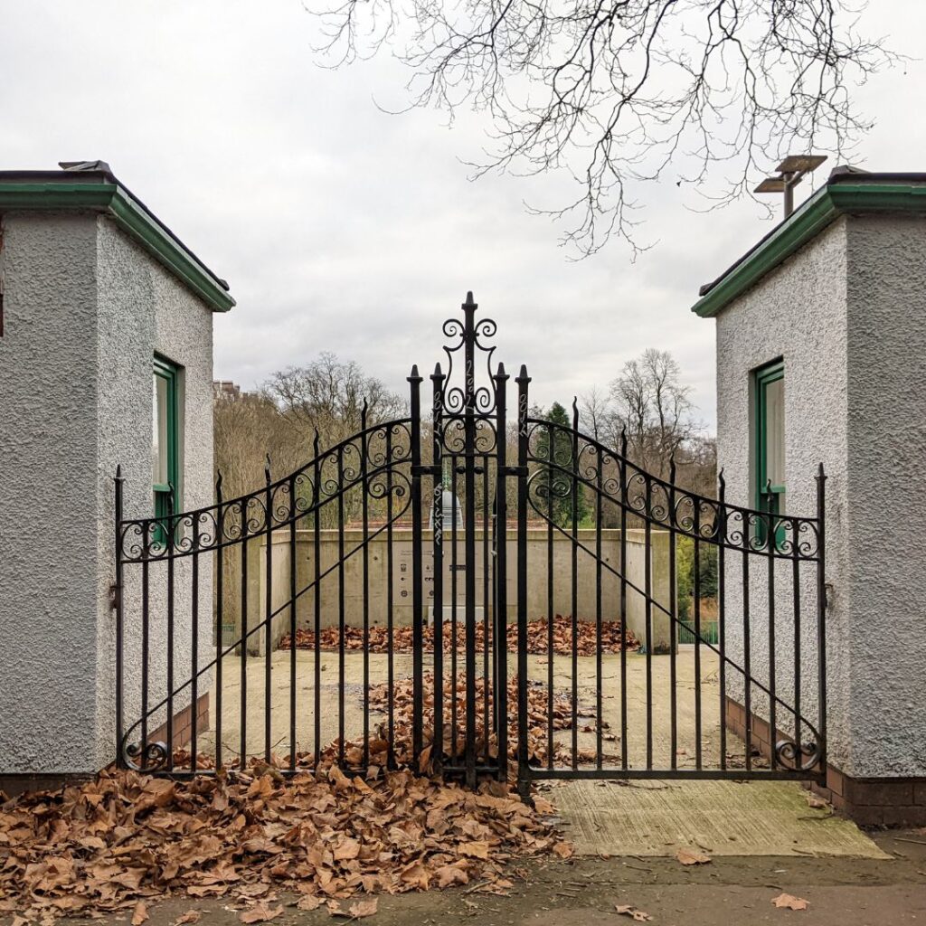A decorative black iron gate stands closed between two white buildings, with fallen brown leaves scattered on the ground and bare trees in the background.