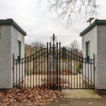 Black wrought iron gates are closed between two small white buildings, with dry leaves scattered on the ground and trees visible in the background under a cloudy sky.