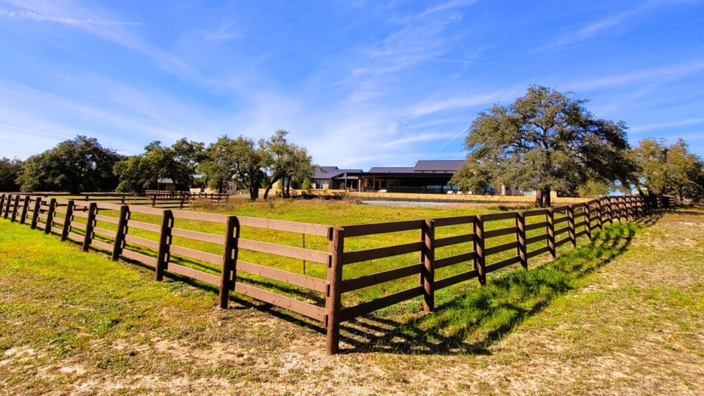 A wooden fence encloses a grassy area on a ranch, with a house and several trees visible in the background under a clear blue sky.