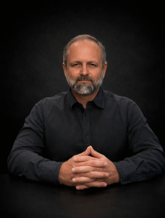 A middle-aged man with a beard, wearing a dark shirt, sits at a table with his hands clasped against a dark background.