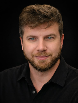 A man with light brown hair, beard, and blue eyes is wearing a black collared shirt and looking directly at the camera against a dark background.
