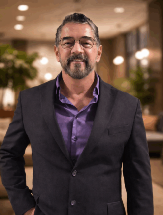 A man with glasses and a beard, wearing a dark blazer over a purple shirt, stands indoors with blurred lights and plants in the background.