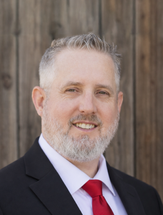 A man with short gray hair and a beard, wearing a black suit, white shirt, and red tie, stands in front of a wooden background.