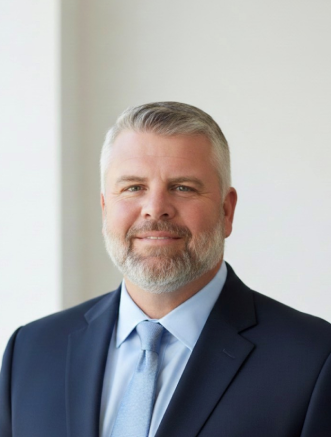 A man with short gray hair and a trimmed beard wears a dark suit, light blue shirt, and matching tie, standing against a plain light background.