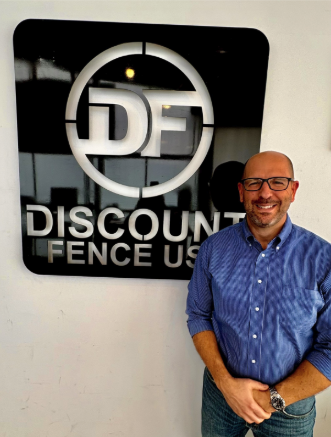 A man wearing glasses and a blue shirt stands and smiles next to a wall sign that reads "DF Discount Fence USA.