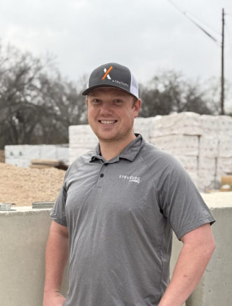 A man in a gray polo shirt and branded cap stands outdoors at a construction site with building materials in the background.
