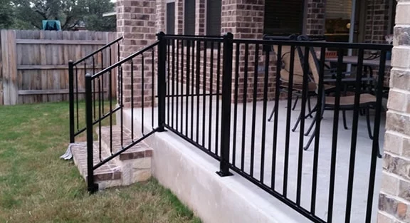 A black metal railing lines the edge of a raised concrete patio with steps, attached to a brick house. Patio furniture is visible in the background.