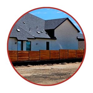 A white modern house with a dark roof and several windows stands behind a wooden fence, with dirt ground in the foreground.