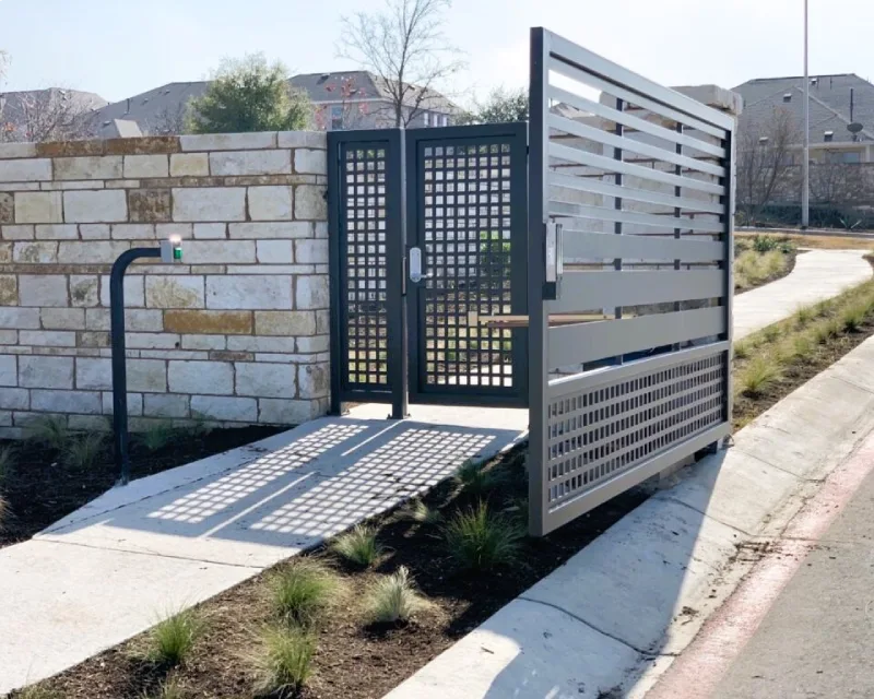 A sliding security gate is open beside a stone wall, blocking the sidewalk, while an access control panel and signs of recent deck installation are visible nearby.