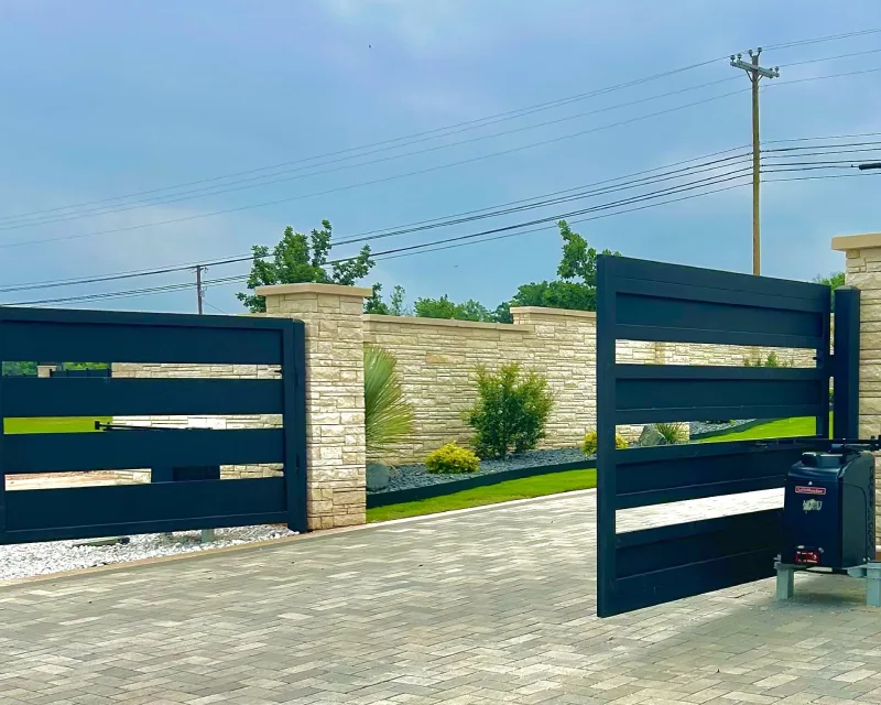 A modern black metal gate is partially open, revealing a paved driveway, stone wall, green lawn, and small plants—perfect for enhancing curb appeal alongside a new deck installation under a cloudy sky.