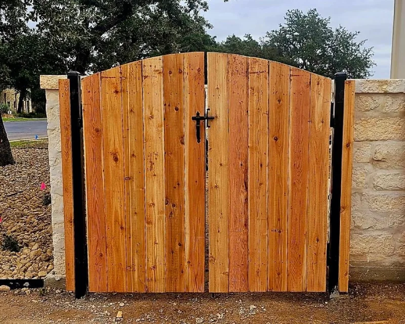 A wooden gate with a curved top and black metal handle, set between stone pillars and a rock landscaping area, offers inviting access to your outdoor space—perfect for complementing any deck installation.