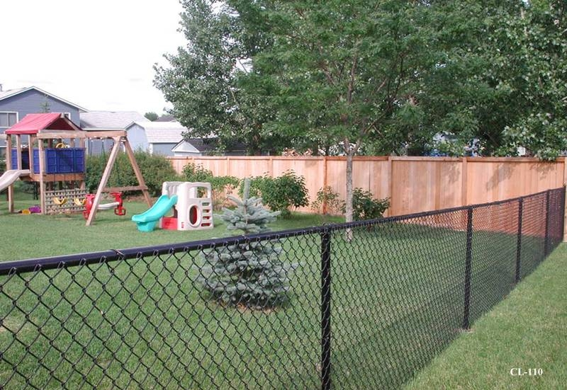 A backyard with a chain-link fence, a small evergreen tree, a wooden playset, a plastic playhouse, and a tall wooden privacy fence in the background.