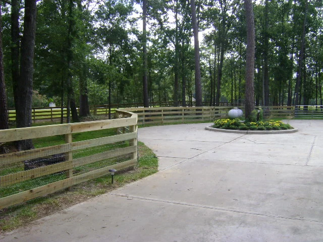 Concrete driveway curves around a circular flower bed with a fountain, surrounded by a wooden fence and tall trees in the background.