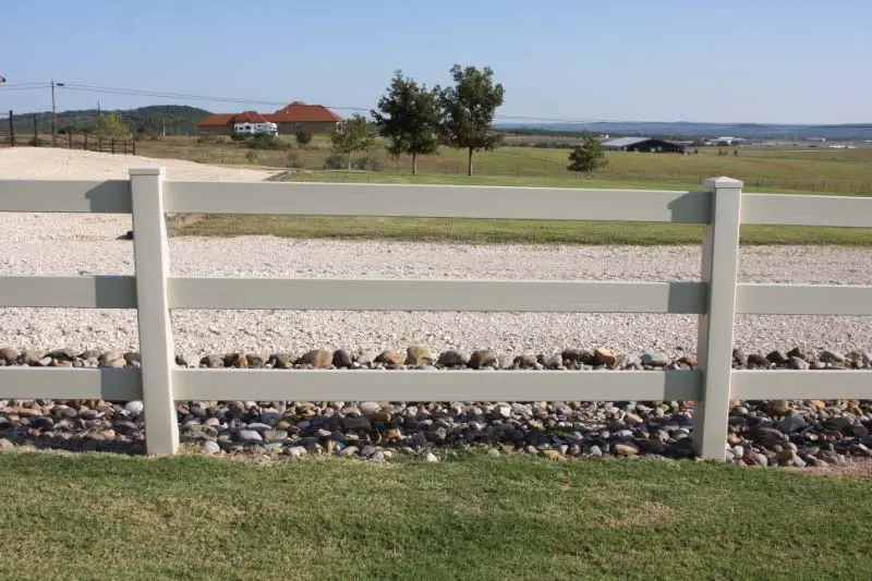 A white three-rail fence runs alongside a gravel path with rocks beneath, grassy fields, trees, and houses in the background under a clear sky.
